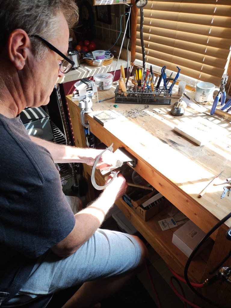 A man working on a project at a wooden workbench, surrounded by various tools and materials, with sunlight streaming in through a window.