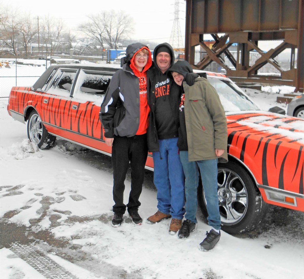 Three people smiling together in winter clothing, standing in front of a car decorated with a tiger stripe design, in a snowy outdoor setting.